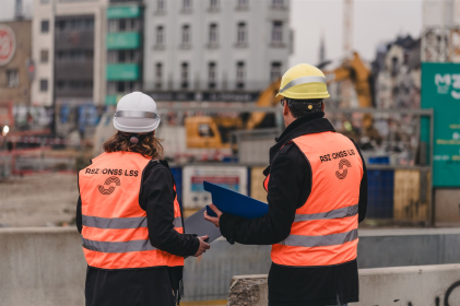 Twee personen met veiligheidshelmen en fluohesjes overleggen op een bouwplaats.